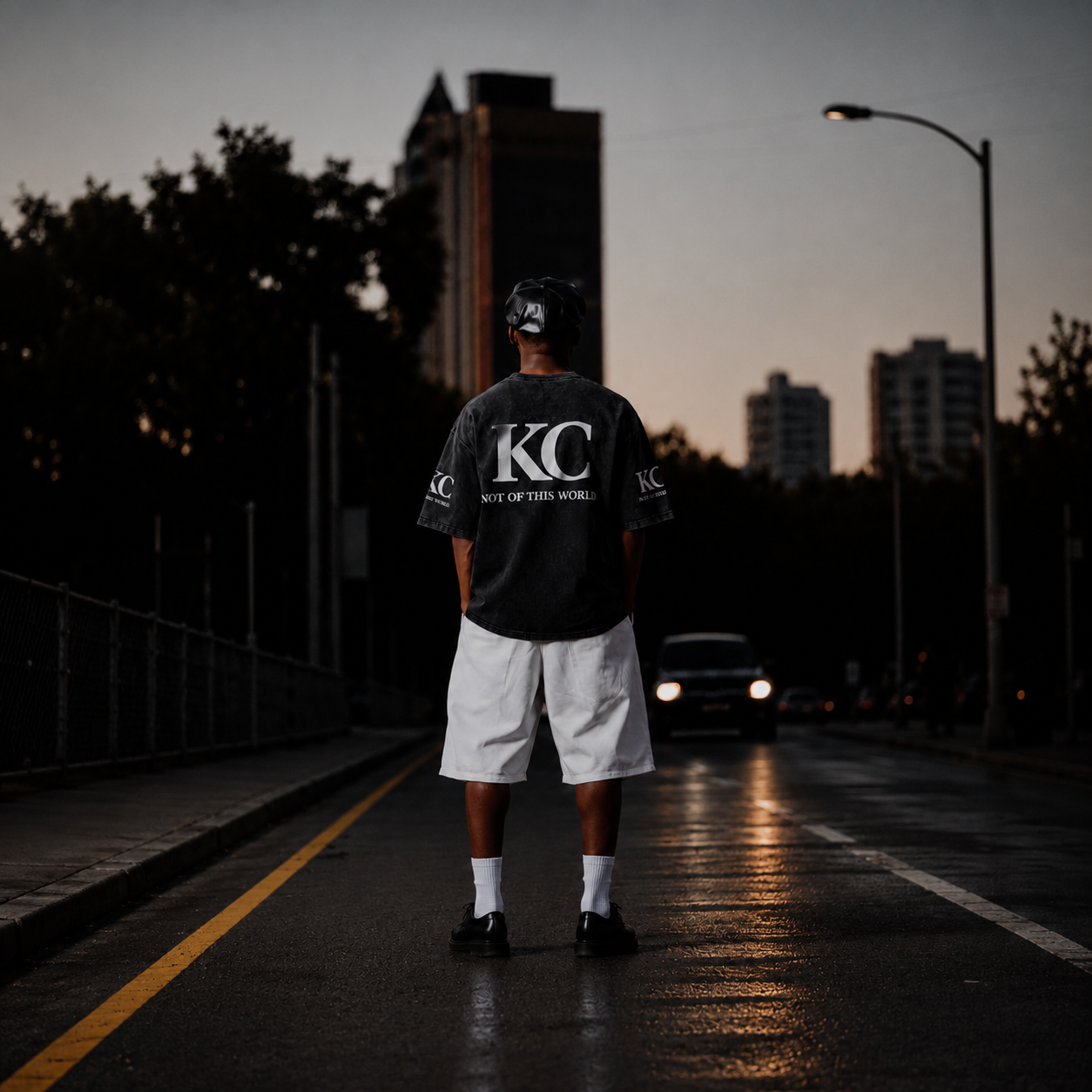 A man stands on a wet city street at dusk, viewed from behind, wearing a dark gray t-shirt with a large white 'KC' logo on the back, white shorts, white socks, and black sneakers.