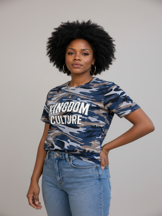 A woman with an afro hairstyle wearing a blue, gray, and beige camouflage t-shirt with 'KINGDOM CULTURE' printed in white across the chest, paired with light blue jeans, against a plain light gray background.