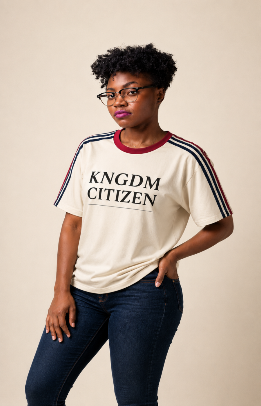 A woman wearing a cream-colored graphic t-shirt with 'KNGDM CITIZEN' printed on the chest, featuring red and navy stripes on the shoulders, paired with dark blue jeans, against a solid beige background.